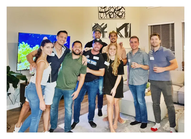 group of people smiling indoors with a big TV on the background