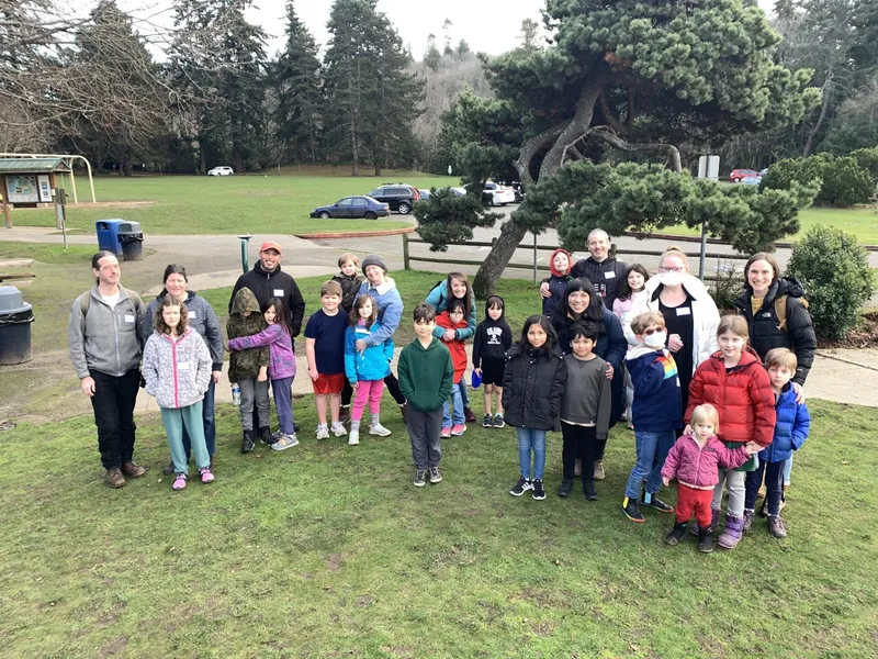A group of people and children outdoors with sweaters on looking at the camera, a park, and trees is shown.